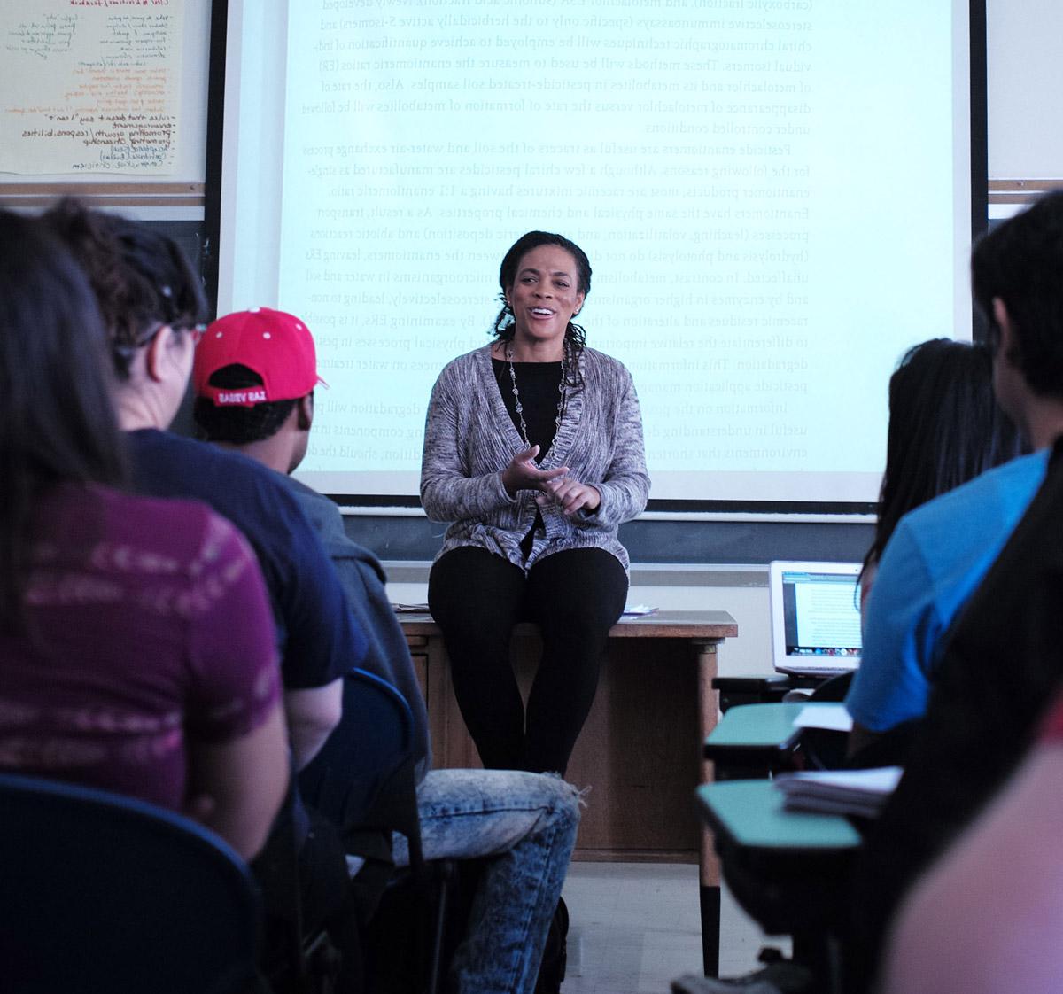 A faculty member sitting on a desk speaking to their class.